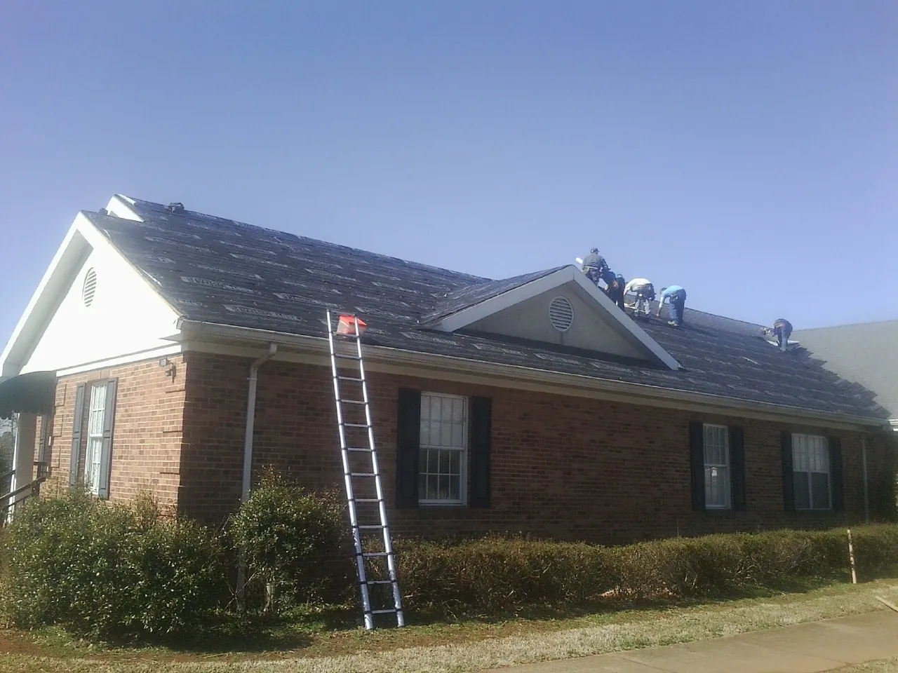 Crew tearing shingles off a brick ranch roof with a ladder set — Emergency Roof Repair work by our crew in Lake Park